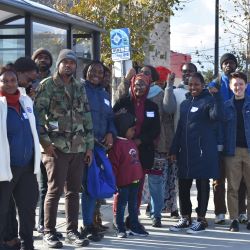 A group of 12 people stand waving with a bus stop behind them