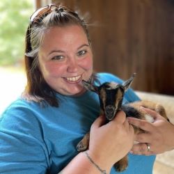 A person with brown hair and a blue shirt smiles at the camera while holding a small baby goat