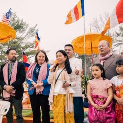 A person with a microphone speaks outside while 5 people stand behind them, with flags and orange umbrellas in the background