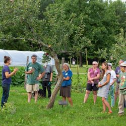 A group of people listen to a speaker outside, with trees and a greenhouse in the background