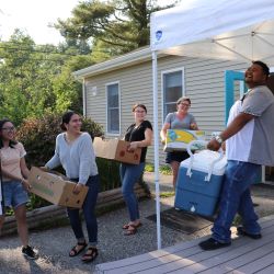 Five people smile and laugh as they bring boxes and a cooler out of a tan building