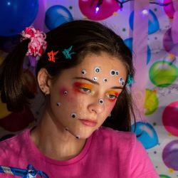 A young person with a pink sweatshirt, bright eye makeup, and googly eyes on their face stares into the distance with tears on their face in front of an image of birthday balloons and a photo of their face with a wide open mouth