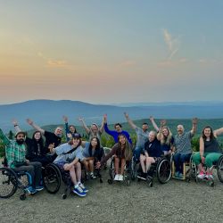 A group of smiling people who use wheelchairs raise their arms in front of a beautiful sunset at the top of a mountain
