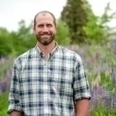 Bill has a beard, brown hair, is wearing a white and green plaid collared shirt and standing in a field of lupine flowers