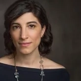 Woman with black hair pulled back, a black top, necklance and a dark background