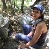 Archana has curly dark hair and sits on a rock on a trail amid trees wearing a billed cap and smiling
