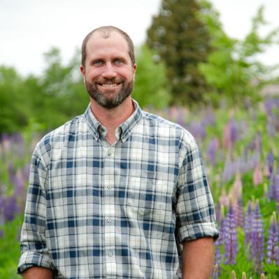 Bill has a beard, brown hair, is wearing a white and green plaid collared shirt and standing in a field of lupine flowers