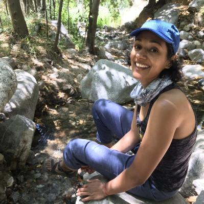 Archana has curly dark hair and sits on a rock on a trail amid trees wearing a billed cap and smiling